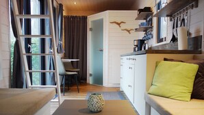 A cozy living area with a sofa, a coffee table, a ladder, and a kitchen area with shelves and a window.