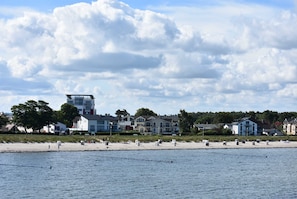Ein Strand mit weißem Sand und eine Reihe von Liegestühlen. Im Hintergrund sind Häuser und ein Gebäude mit flachem Dach zu sehen.