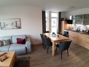 Ett modernt vardagsrum med en soffa, ett matbord med stolar, en köksdel med skåp och vitvaror samt ett stort fönster med gardiner.