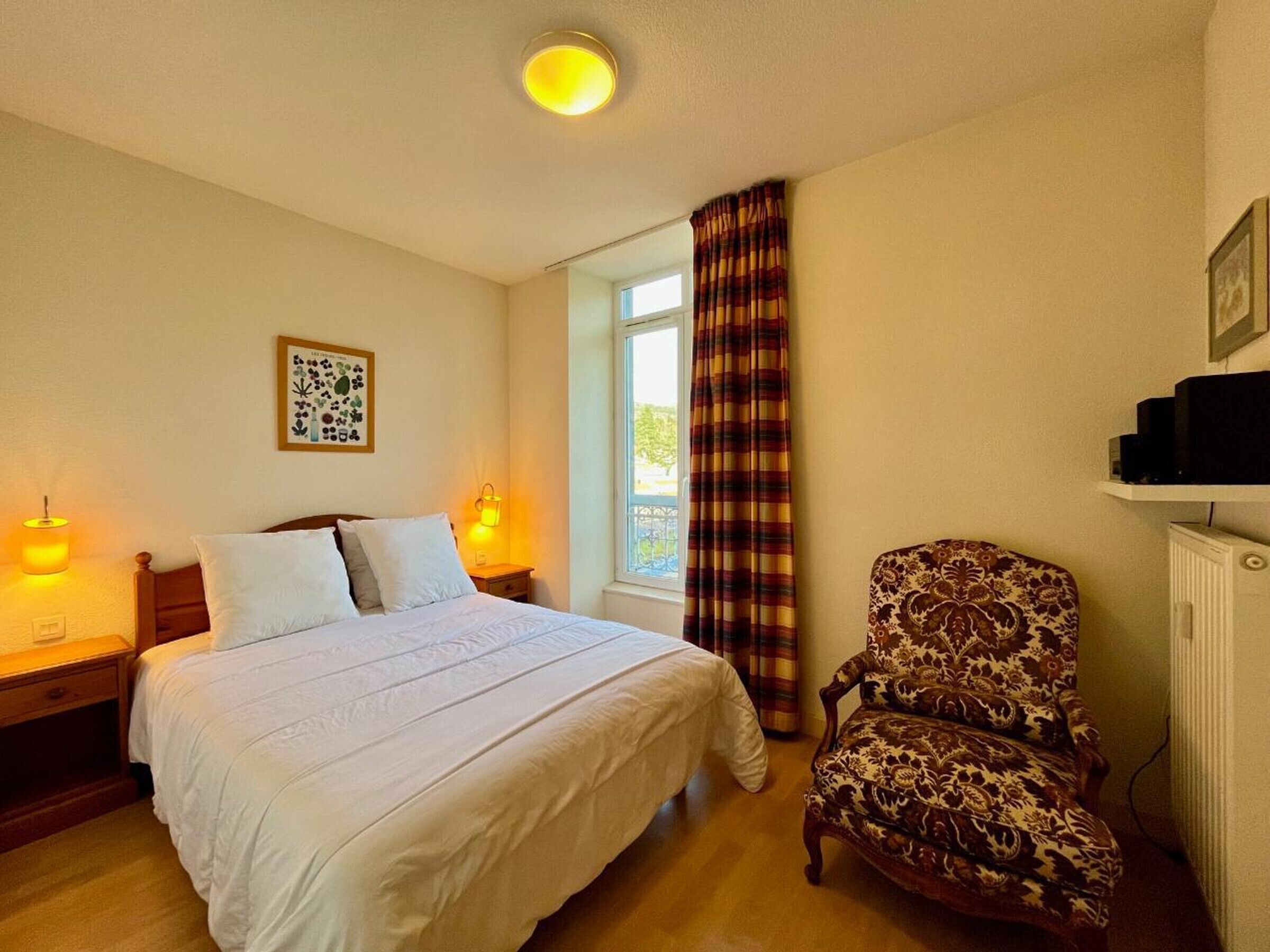 Une chambre à coucher comprenant un lit, une chaise, une fenêtre avec des rideaux, un tableau accroché au mur et un radiateur.