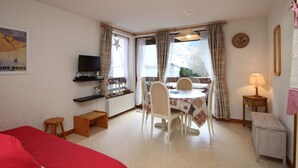 A cozy room with a bed, a dining table with chairs, a TV on a shelf, and a window with curtains.