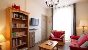 A living room with a wooden bookshelf, a television mounted on the wall, a red sofa, a wooden coffee table, and a chandelier.