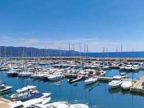 Un port de plaisance où de nombreux bateaux sont amarrés, une jetée qui s’avance dans l’eau et un ciel bleu azur.