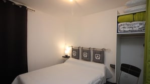 Uma cama de solteiro com colcha branca, uma mesa de cabeceira com abajur, uma cadeira e uma estante com cobertores dobrados.