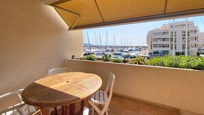 Varanda com mesa e cadeiras, vista para uma marina com barcos e um edifício moderno ao fundo.