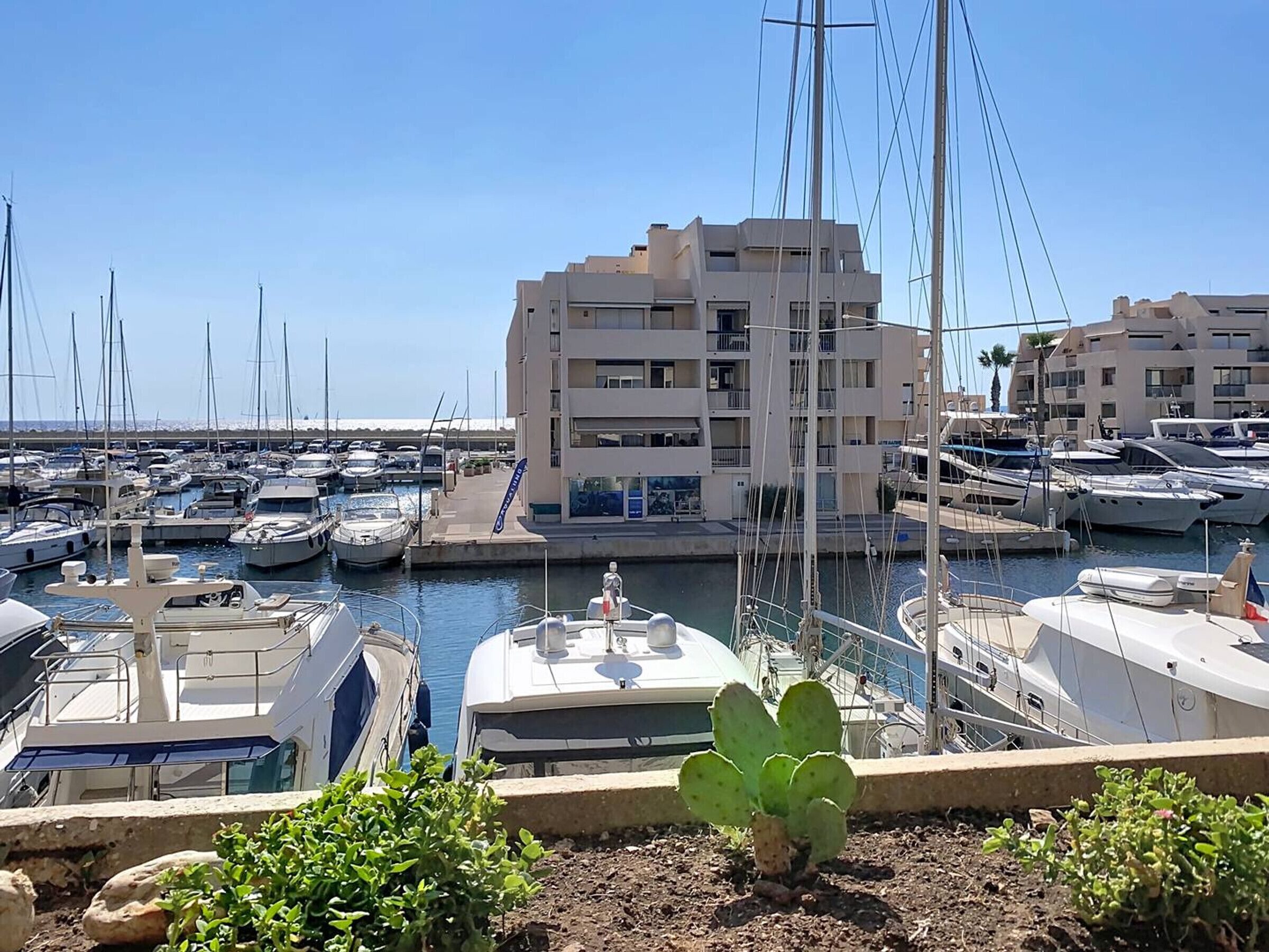 A marina with multiple boats docked, a building with balconies, and a clear sky.