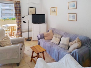 Ein Wohnzimmer mit einer Couch, Sesseln, einem kleinen Holztisch, einem Fernseher auf einem Schrank und gerahmten Bildern an der Wand.