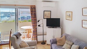 Uma sala de estar com sofá, uma poltrona, uma mesinha, uma TV sobre um móvel e uma varanda com vista para barcos e edifícios.