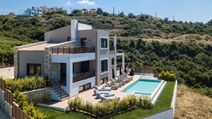Una casa moderna de dos pisos con piscina, rodeada de vegetación y colinas.