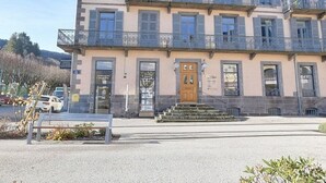 A multi-story building with a central entrance, a staircase leading to the entrance, and a small outdoor seating area. - T3 Apartment in La Bourboule Center with Balcony and Small Dog Allowed (La Bourboule)