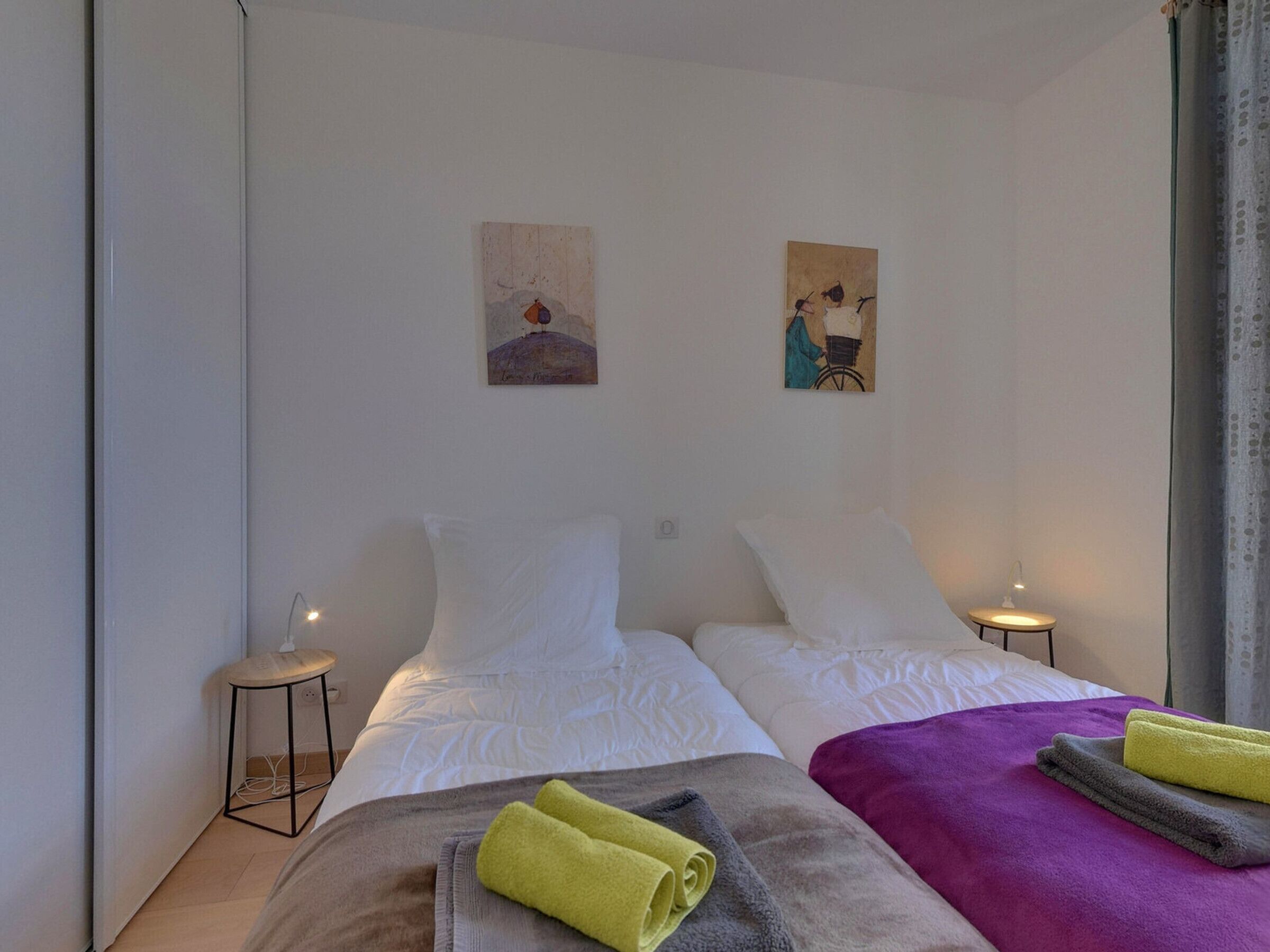 Een hotelkamer met twee bedden, witte lakens, een paarse deken en twee schilderijen aan de muur.