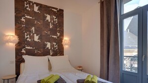 A hotel room with a bed, two pillows, a nightstand, a small stool, and a window with curtains. - T3 Apartment in La Bourboule Center with Balcony and Small Dog Allowed (La Bourboule)