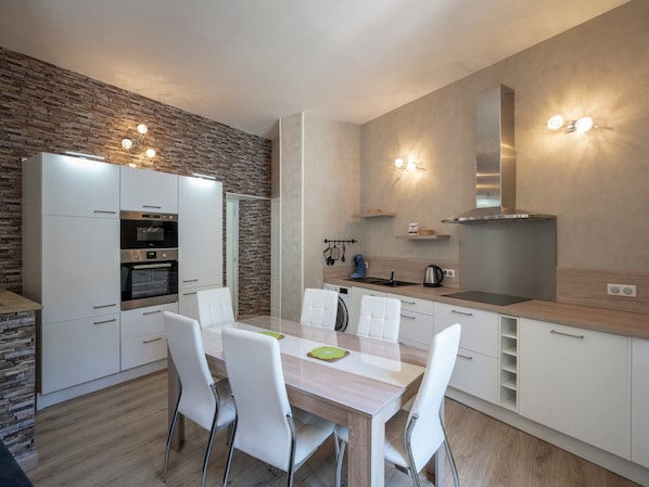 A modern kitchen with white cabinetry, a stone accent wall, stainless steel appliances, and a dining area with white chairs. - MONT DORE - Charming T2 Plus with WIFI, Sleeps 6 (Mont-Dore)