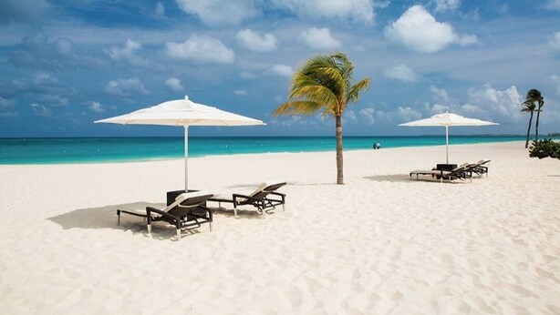 Een strand met wit zand, twee witte parasols en ligstoelen.