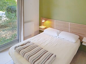 A bedroom with a bed, a nightstand, a lamp, and a window with a view of greenery.