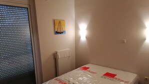 A room with a bed covered in protective plastic, a radiator, a window with blinds, and a wall-mounted light. - Beach House (artemida)