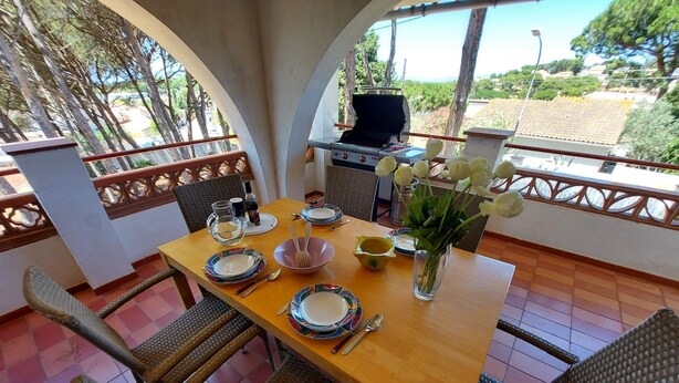 A dining table set with plates, cutlery, and a vase of flowers, surrounded by wicker chairs, with a grill and outdoor view. - Villa Catabella with unlimited WiFi internet next to the beaches and the village (L'Escala)