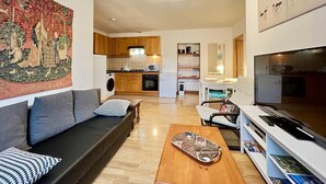 A modern living room with a sofa, coffee table, and TV. There is a kitchen in the background with wooden cabinets and a microwave. - La Bourboule: Comfortable T3, Parking, WiFi (La Bourboule)