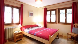 A bedroom with a wooden bed, two bedside tables, a nightstand, a chair, and a window with curtains. - Chalet 6 rooms, sleeps 8, near center, WIFI (Mont-Dore)