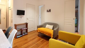 Una habitación pequeña y bien iluminada con un sofá, un sillón amarillo, una mesita de madera y un televisor montado en la pared.