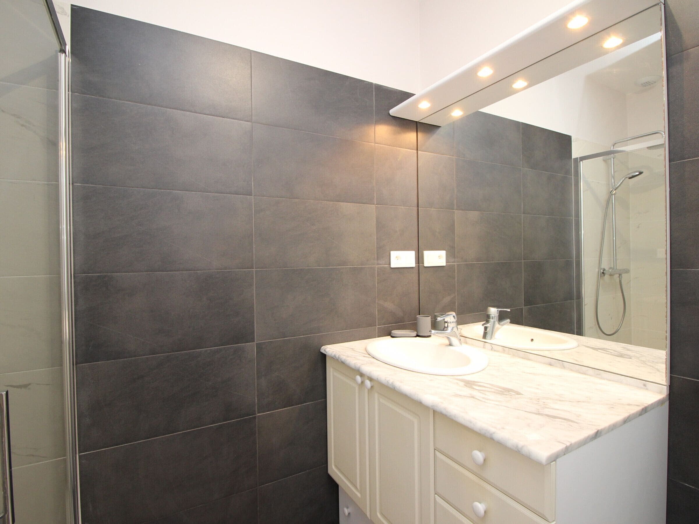 A modern bathroom with dark tiles, a glass shower enclosure, and a marble countertop.