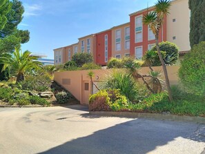 A multi-story building with a beige and red facade, surrounded by lush greenery and palm trees. - Spacious 2-Bed Cabin Apartment Near Beach & Shops, Large Loggia, Garage Included (Bormes-les-Mimosas)