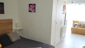 Ein Schlafzimmer mit einem Bett, einem Nachttisch mit Lampe, einem Kleiderschrank und Blick auf einen Wohnbereich mit einer Couch.