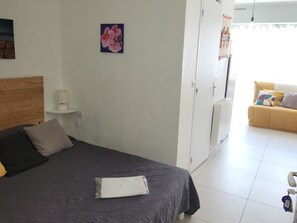 A bedroom with a bed, a bedside table with a lamp, a wardrobe, and a view of a living area with a sofa.