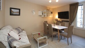 A modern living room with a sofa, a glass coffee table, a dining table with chairs, a TV mounted on the wall, and a window with curtains. - Mont-Dore: Central T3 Apartment with Wifi (Mont-Dore)