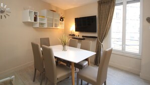 A dining area with a white table and four chairs, a TV mounted on the wall, and a window with curtains. - Mont-Dore: Central T3 Apartment with Wifi (Mont-Dore)