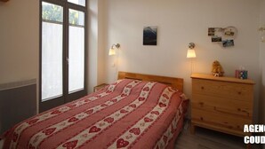 Ein Schlafzimmer mit großem Fenster, einem Bett mit decke mit Herzmuster, Nachttischlampen, einer Kommode und einer Wand mit Fotos und einem kleinen Gemälde.