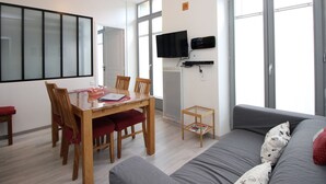 Ein modernes Wohnzimmer mit Esstisch, Stühlen, einem grauen Sofa, einem flachbildfernseher an der Wand und großen Fenstern, die für natürliches Licht sorgen.