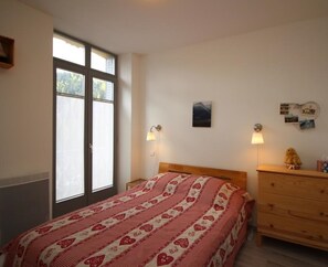 Un dormitorio con un ventanal grande, una cama con manta estampada de corazones, lámparas de noche, un aparador y una pared con fotografías y un cuadro pequeño.