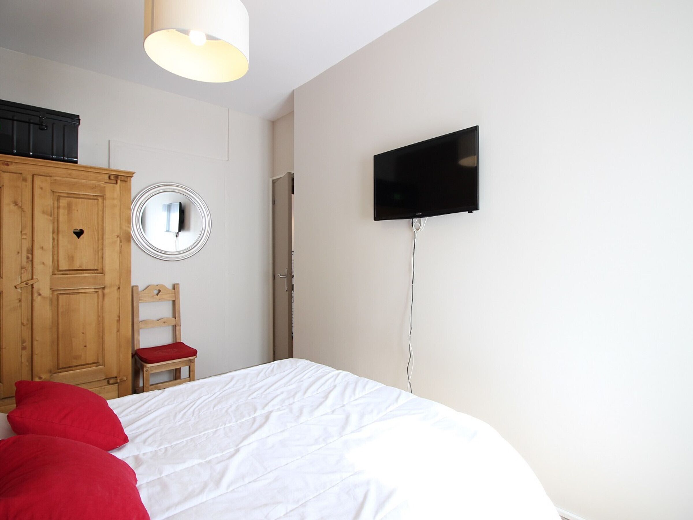 Une chambre à coucher avec un lit, un téléviseur fixé au mur, une armoire en bois, un miroir et une chaise rouge.