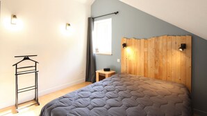 Un dormitorio con techo inclinado, una cama con colcha gris, cabecera de madera, mesita de noche de madera, perchero negro y una ventana con cortinas.