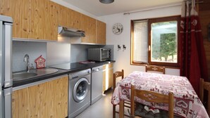 A compact kitchen with wooden cabinets, a washing machine, a microwave, and a table with a patterned tablecloth. - Charming T2 with balcony in Mont Dore, parking included (Mont-Dore)