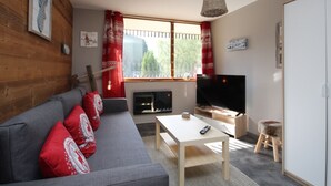 A living room with a grey sofa, red cushions, a TV, a fireplace, and a window with red curtains. - Charming T2 with balcony in Mont Dore, parking included (Mont-Dore)