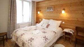 Une chambre à coucher avec une tête de lit en bois, un lit recouvert d’une literie blanche et d’une couverture à motifs croisés, deux lampes de chevet et une fenêtre avec des rideaux.