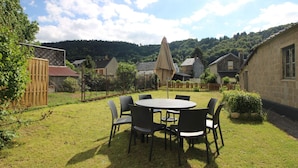 A round table with chairs in a grassy backyard, surrounded by stone buildings and a wooden fence. - Mont-Dore, renovated 2-bedroom with garden, parking, wifi (Mont-Dore)