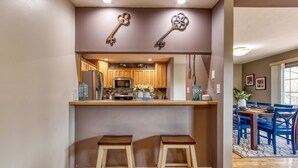 Une cuisine avec des meubles en bois, un réfrigérateur en acier inoxydable et un four à micro-ondes. On y trouve deux tabourets placés sous un comptoir, et une décoration en forme de clé est accrochée au-dessus du comptoir.