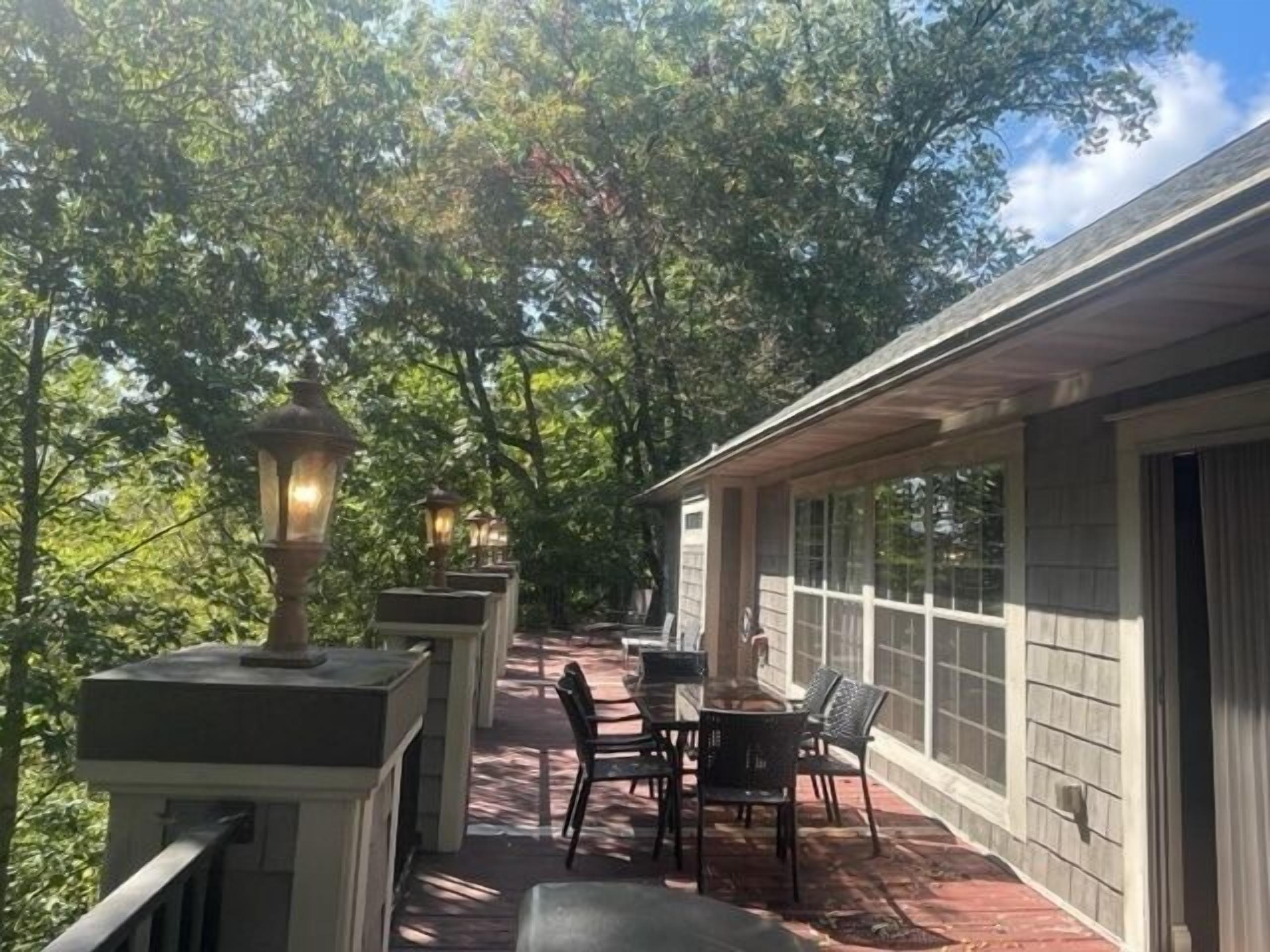 A covered outdoor patio with a table and chairs, surrounded by trees and a building with large windows.