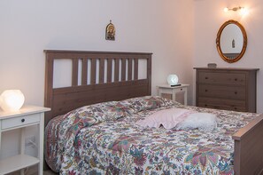A bedroom with a wooden headboard, a floral bedspread, a bedside table with a lamp, a dresser with drawers, and a wall-mounted mirror.