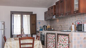 A compact kitchen with wooden cabinets, a dining table with chairs, a refrigerator, a stove, and a microwave. - Beautiful 1-bed House in Avola (Avola)