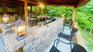 Un área de cocina al aire libre con paredes de piedra, asientos de bar y vistas a follaje exuberante.