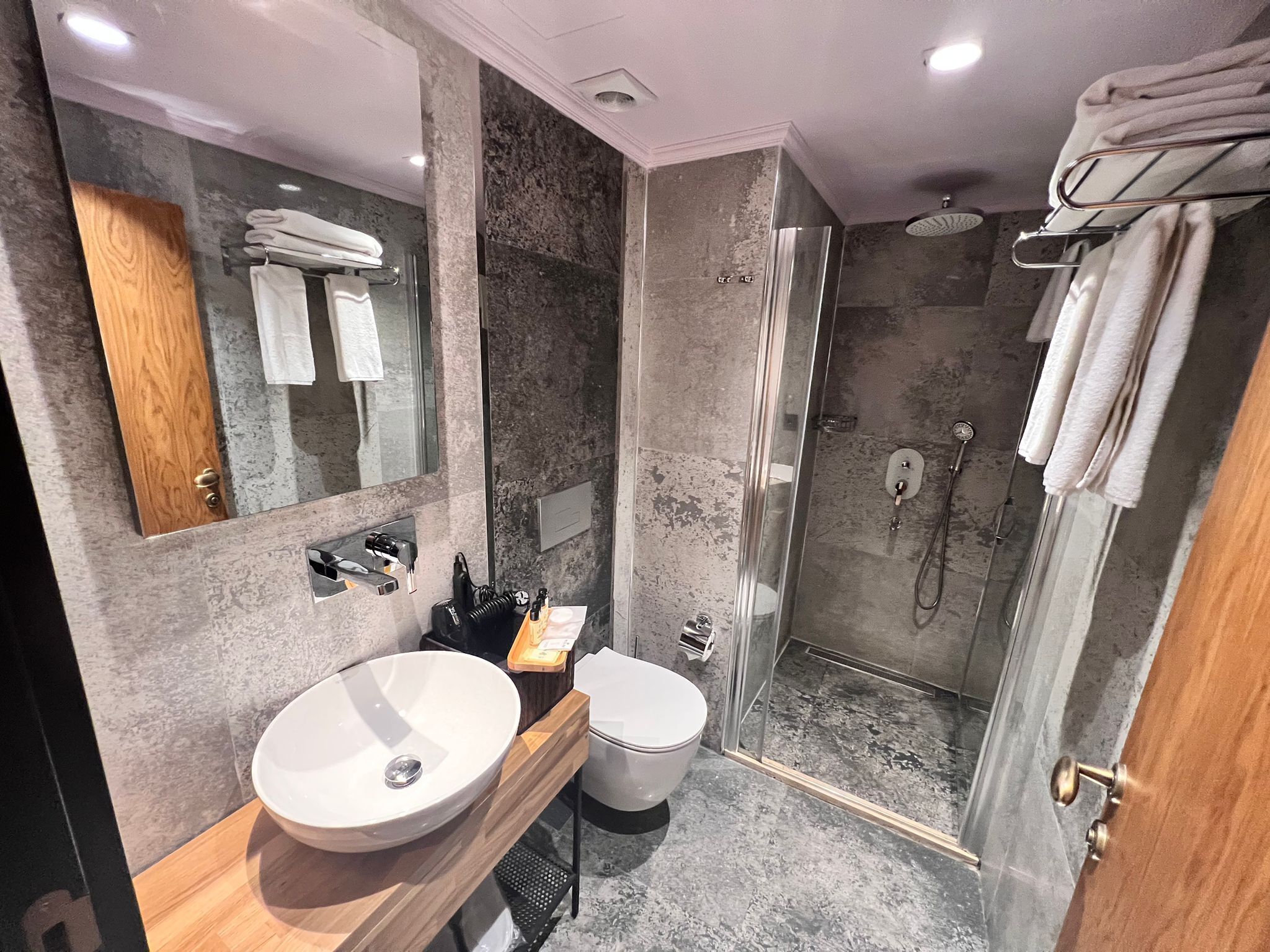 A modern bathroom with a large mirror, a wooden vanity, a white sink, a toilet, and a glass-enclosed shower area.