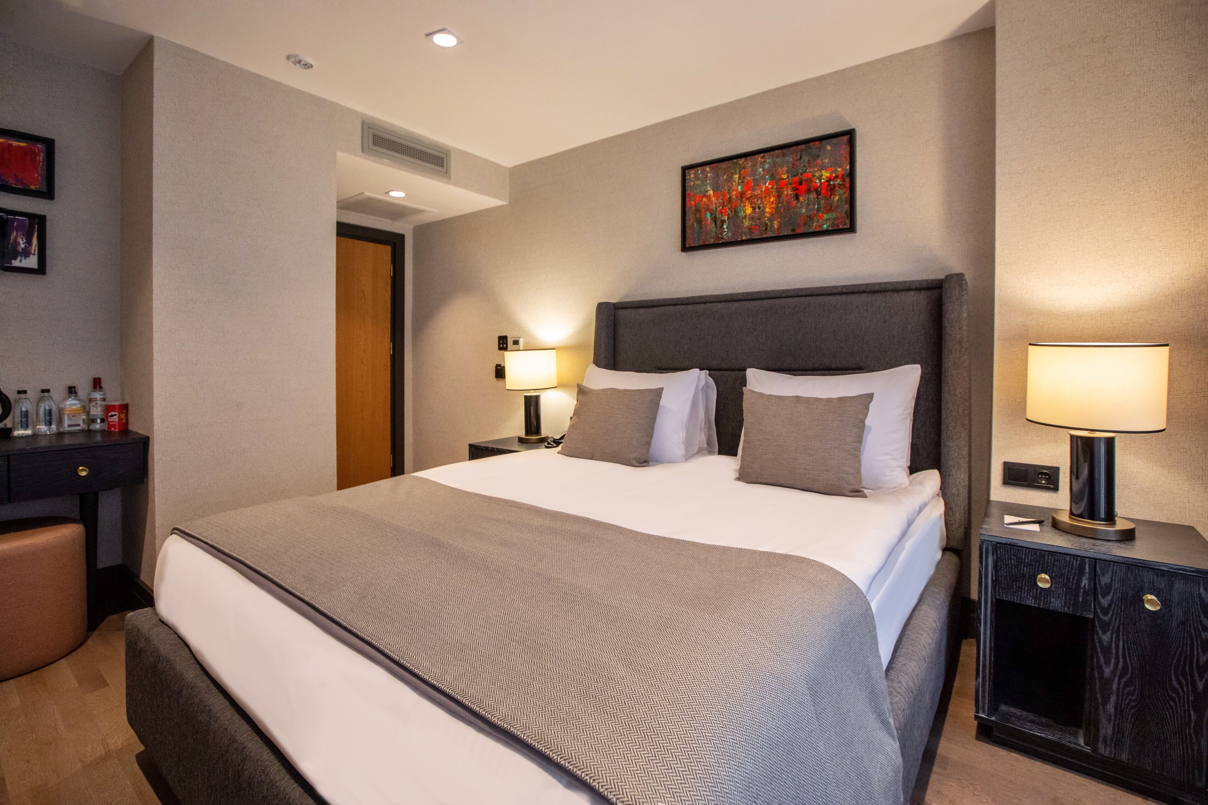 A hotel room with a large bed, two bedside tables, a desk with a chair, and a painting on the wall.