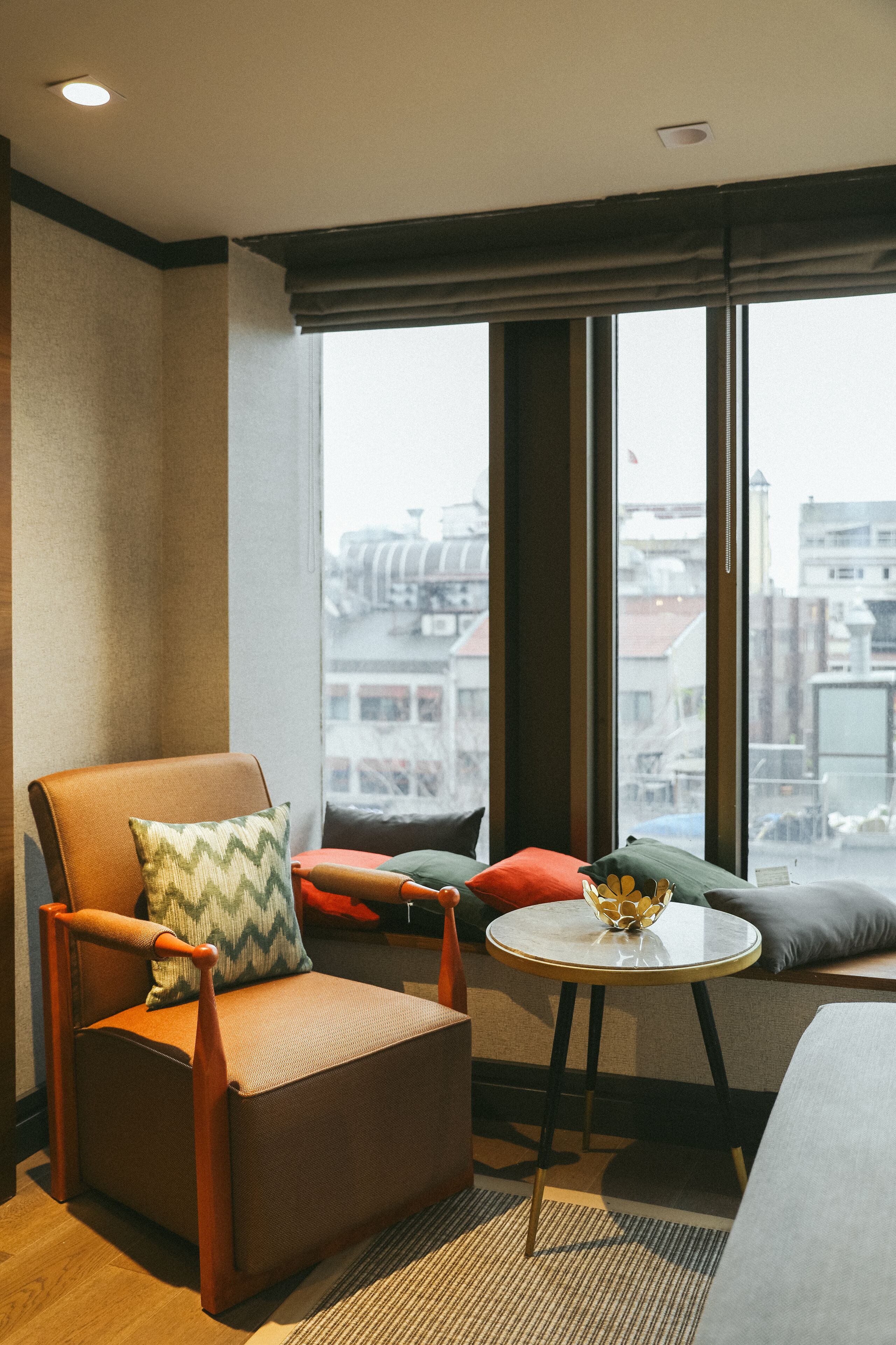 A modern hotel room with a large window, a comfortable armchair, a small round table, and a sofa with multiple cushions.