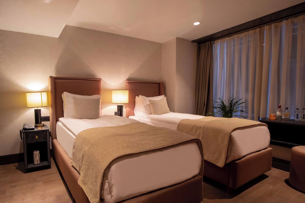 A hotel room with two beds, a nightstand with a lamp, a small table with toiletries, and a large window with curtains.