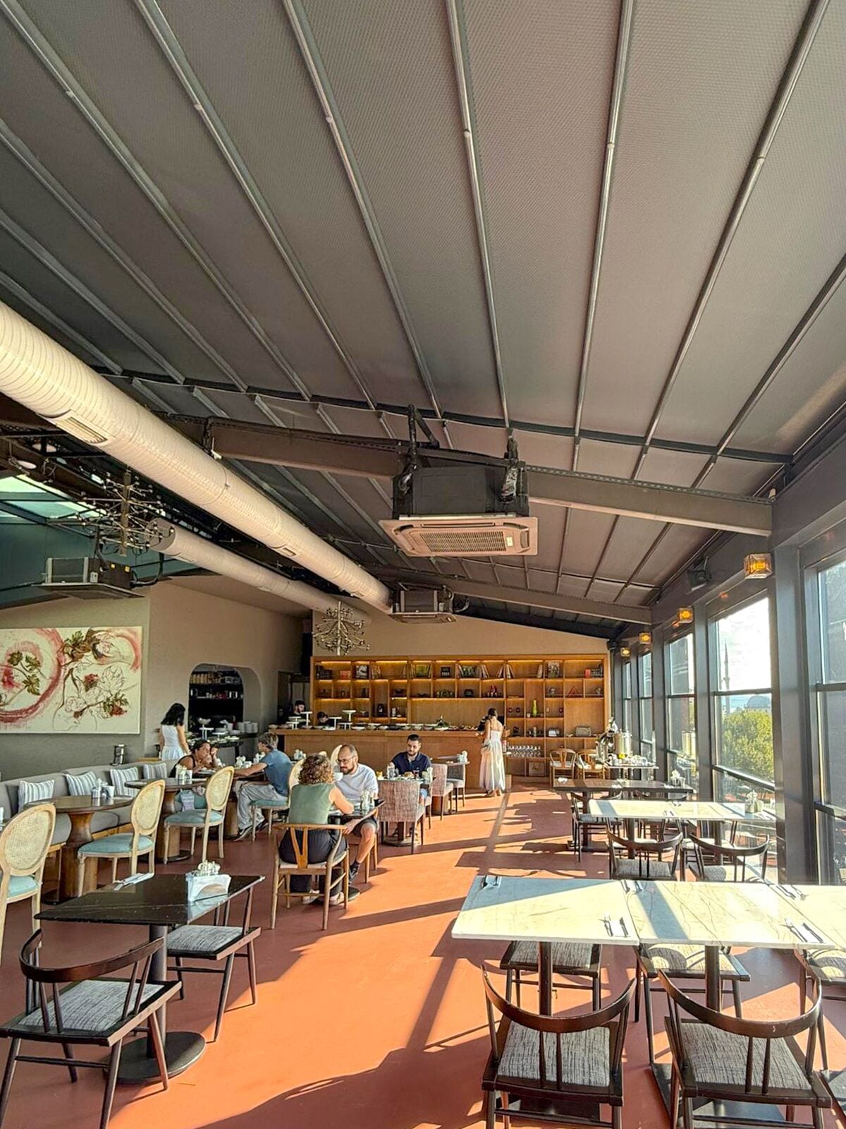 Uma área de refeições interna com mesas e cadeiras, um bar e uma janela ampla que proporciona luz natural.