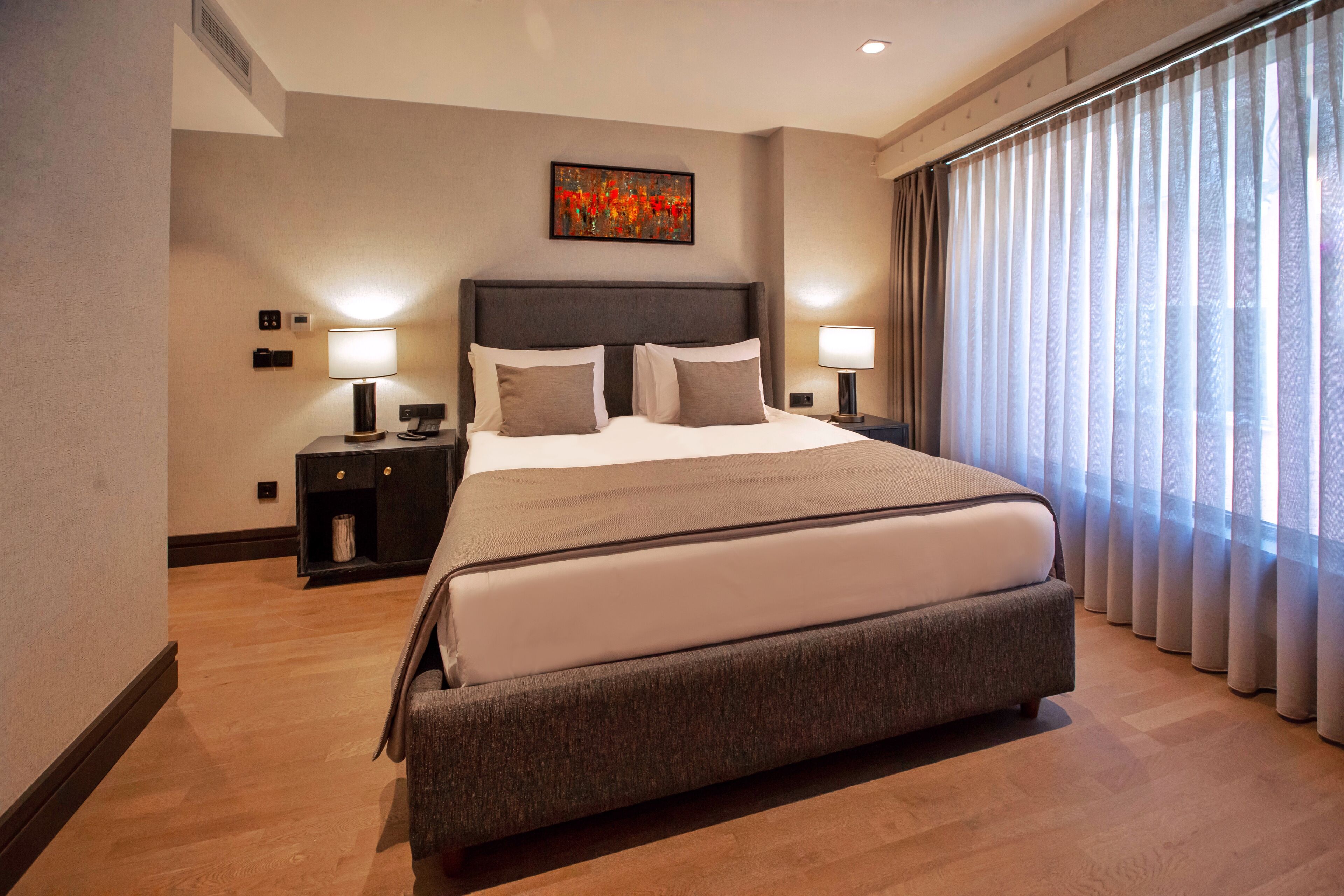 Habitación de hotel con una cama grande, dos mesitas de noche, un aparador, un cuadro en la pared y una ventana con cortinas.